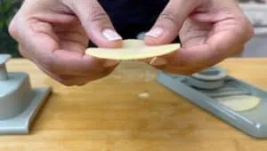 Close-up of hands holding a thinly sliced potato over a mandoline slicer on a wooden cutting board.